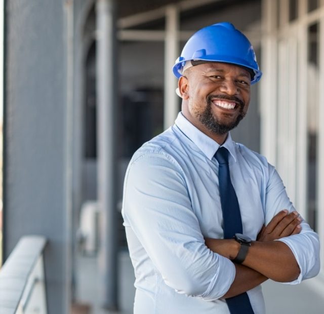 Portrait of african american man architect at building site with folded arms looking at camera. Confident construction manager in formal clothing wearing blue hardhat. Successful mature civil engineer at construction site with copy space.